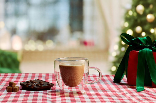 Cup of sweet cocoa on tablecloth and dark chocolate with nuts. Concept of hot drinks, taste, winter season, coziness, warm atmosphere. Beautiful holiday background with lights and Christmas tree