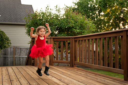 An adorable two year old is happily dancing the evening away on her back porch. She's wearing a bright red ballerina dress with black rain boots. She is ethnically ambiguous.