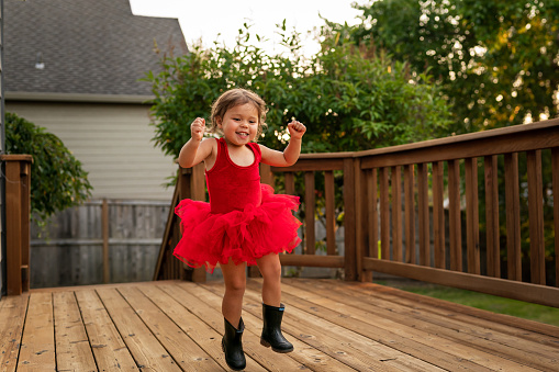 An adorable two year old is happily dancing the evening away on her back porch. She's wearing a bright red ballerina dress with black rain boots. She is ethnically ambiguous.
