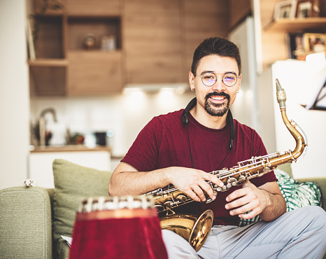Portrait of Young male musician practising saxophone at home. He is barefoot and wearing eyeglasses, dressed in casual clothes. Interior of small apartment during day time.