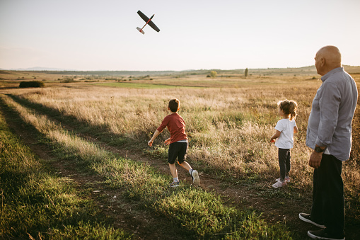 Grandfather with his grandson and granddaughter having fun on a meadow in sunset. Young boy is throwing airplane toy.