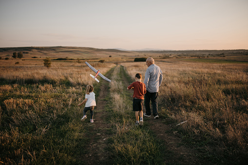 Grandfather with his grandson and granddaughter having fun on a meadow in sunset. Little girl is throwing airplane toy.