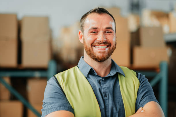 fabrik- oder lagerarbeiter in der versandfabrik glücklich und lächelnd über neue import- und exportfracht. porträt eines jungen logistik- oder supply chain-industriemanagers, der aufgeregt und stolz aussieht - warehouse worker stock-fotos und bilder