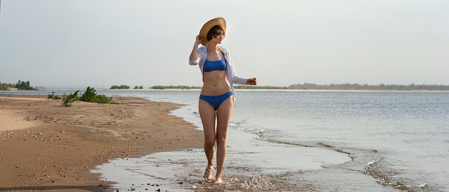 Young woman with perfect body walks and tanning at beach resort, wearing blue bikini, white shirt and straw sun hat. Girl enjoying summer holiday travel. Full length portrait lifestyle outdoor. Banner