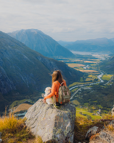 Woman with long hair in orange sitting on the top of the mountain with a cute pug, admiring the view of the mountains, river and the green meadow in More og Romsdal county, Norway