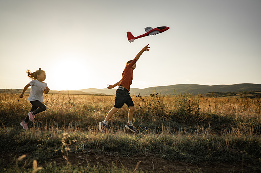 Brother and his little sister playing together on a meadow in sunset. Young boy is throwing airplane toy.