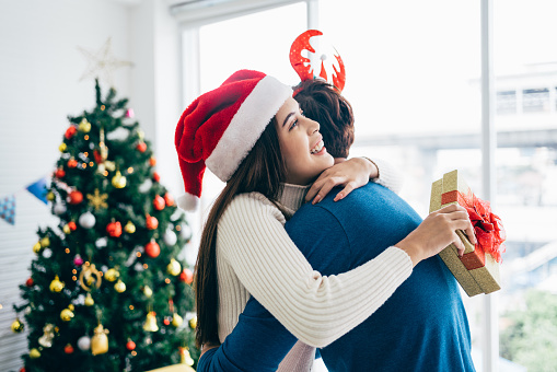 Asian woman with surprise Christmas gift hugging husband at home. Presenting gift as Christmas eve tradition, celebrating New Year. Happy Asian woman receiving congratulations from her boyfriend.