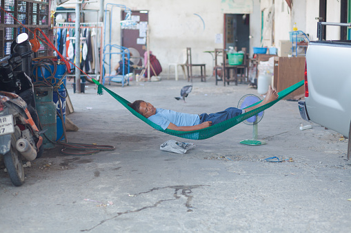 Adult thai man is relaxing and sleeping in hammock in residential district in Bangkok Chatuchak. Hammock is plugged to a pick-up car. Scene is a driveway of a building, Man is wearing casual clothing