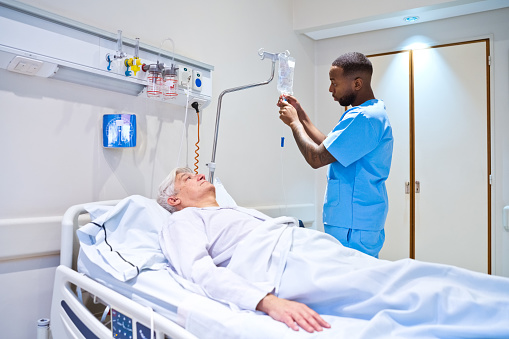 Side view of male nurse checking IV drip while senior patient lying on bed in hospital