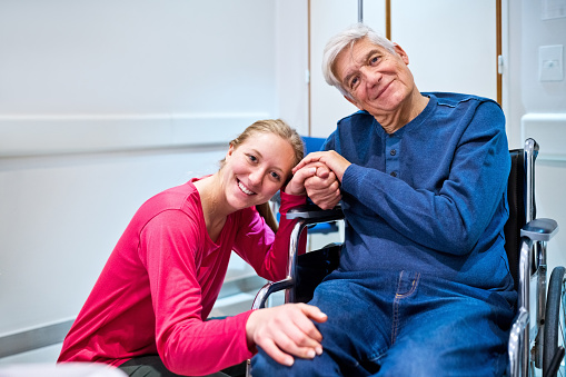 Portrait of smiling granddaughter holding hands of grandfather sitting on wheelchair in hospital