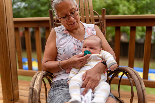 A senior latin woman is sitting on her back porch and relaxing while proudly holding her newborn grandson.
