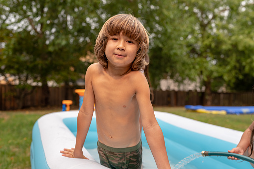 A young latin american boy is playing in his backyard inflateable pool with his siblings.  They are happy and enjoying summer and sunshine. He is looking directly into the camera.