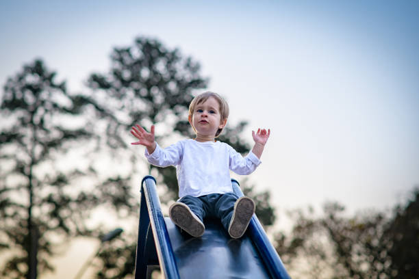 A cheerful toddler boy looking away with arms raised preparing to slide down slide at playground at sunset