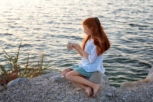 A beautiful girl with long red hair sits on the shore of the sea, ocean, river and holds a large shell in her hands. Concept of tourism, travel, vacation by the ocean in summer.