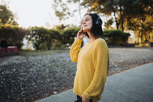 Teenage girl wearing yellow sweater, using headphones for playing music on her smartphone in the park at afternoon