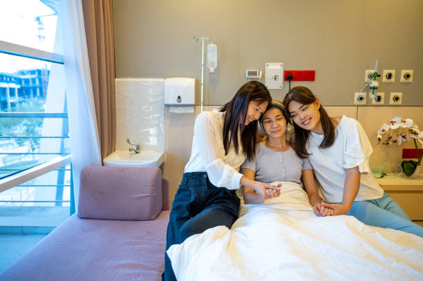 Two Asian female friends visiting patient in hospital ward. An Asian Chinese female patient lying on the bed with her two female friends visiting her in the hospital ward stock photo