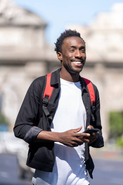 istock Portrait of a happy man walking with red backpack on his back using cell phone 1425661935