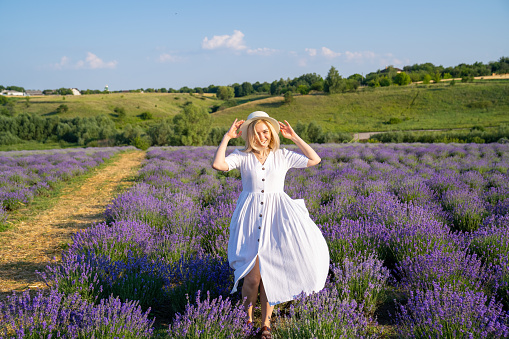 woman model in white dress outfit with hat is standing dancing in lavender field, photo session. Concept of freedom, good mood, enjoying nature, sense of happiness