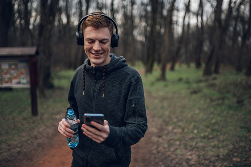 Athletic redhead young man using a smartphone while running outside in the forest path. Listening music on a headphones while running. Athletic and healthy.