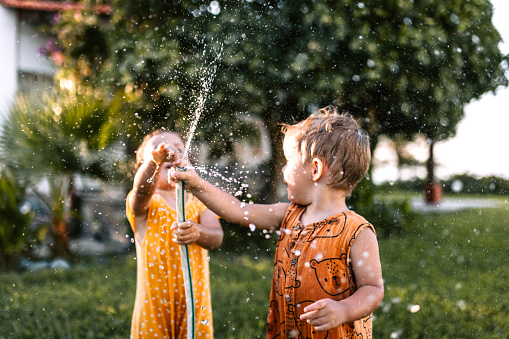 Little brother and sister playing with garden hose in backyard, having fun with spraying water.