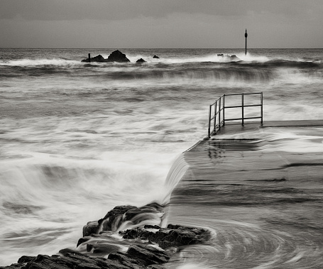 A  black and white image of a cold, wintery evening at the natural bathing pool in Bude, Cornwall, England. The waves swell as they prepare to beat against the shore. They provide a hint of green against the relentless grey of the dusk. The distant rocks with their warning beacons, and the solitary railings remind us that the sea can never really be tamed.