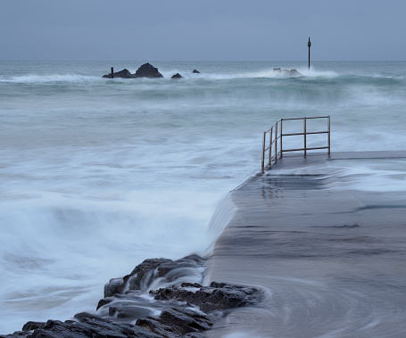 A cold, wintery evening at the natural bathing pool in Bude, Cornwall, England. The waves swell as they prepare to beat against the shore. They provide a hint of green against the relentless grey of the dusk. The distant rocks with their warning beacons, and the solitary railings remind us that the sea can never really be tamed.