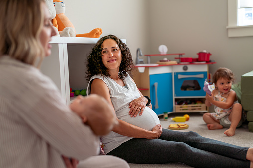 A mixed race pregnant woman sits on the floor at home and smiles while enjoying visiting with a friend who has a newborn baby. The woman's firstborn daughter is in the background playing with toys.