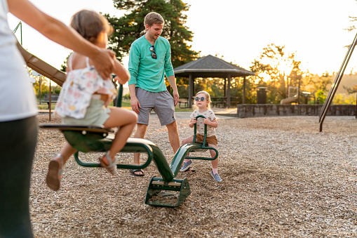 Two young girls ride a seesaw with their mom and dad's help while playing at the park on a warm and sunny evening.