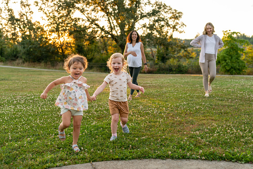 Two toddler best friends are running through a park together holding hands while their mothers walk behind them and talk. One of the moms is of Pacific Islander descent and eight months pregnant.