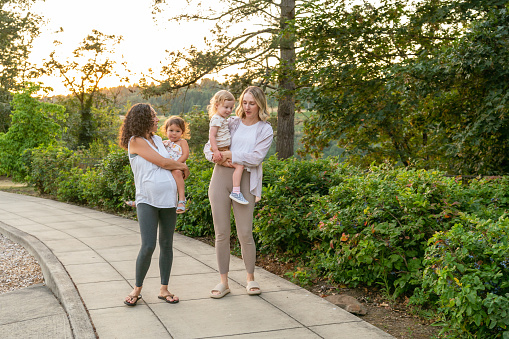 Two young mothers talk happily as they hold their toddler daughters on their hips and walk through the park at sunset. One of the women is eight months pregnant.