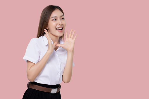 Teen student girl of Asian ethnicity in university uniform open mouths raising hands screaming announcement isolated over pink background.