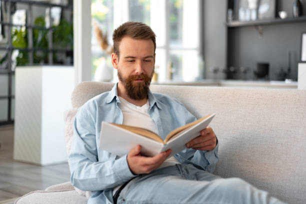istock Young bearded handsome man reading book on couch at home 1425719986