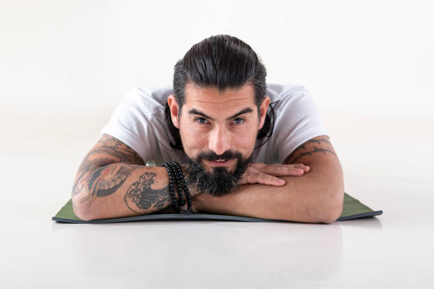 Portrait of a man dressed in white yoga clothes lying on a mat while looking at camera over white background. Portrait of a man dressed in white yoga clothes lying on a mat while looking at camera over white background. Studio shot. man yoga tattoo stock pictures, royalty-free photos & images