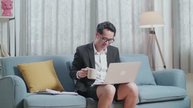 Asian Businessman In Jacket And Shorts Drinking Coffee While Working With A Laptop At Home