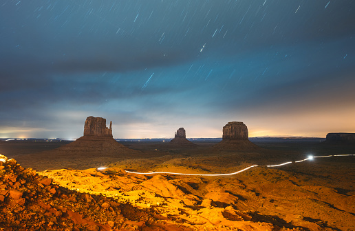 Night sky with stars showing on a dramatic sky above the towering sand stone buttes at Monument Valley. Located on the Arizona and Utah border of the southwest USA. Glowing lights showing from a car passing by on the gravel road.