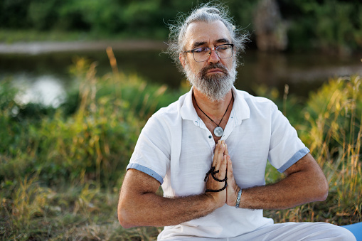 Mature man sitting on exercise mat and meditating in nature by the river