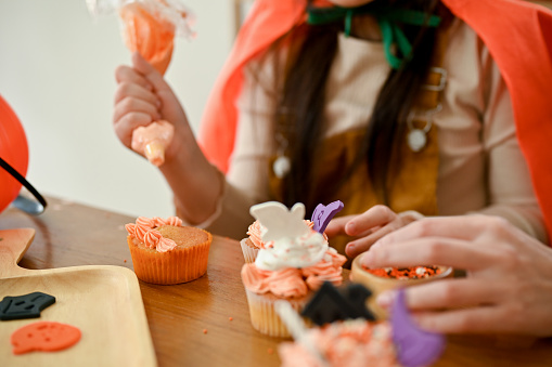 A cute young Asian girl enjoys making a Halloween cupcake with her mom, decorating her cupcake with cream. cropped shot