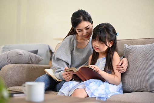 Lovely and relaxed Asian mom reading a book or telling fairy tale story to her cute daughter while relaxing in their living room.