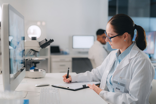 Woman scientist doing research and writing in laboratory for experiment. Analyze results on computer, microscope and clinical diagnosis notes, pharmacy and lab technician or medical assistant.