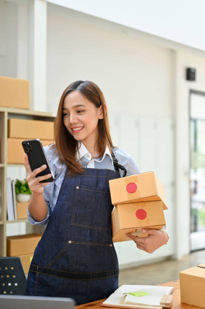 Portrait, Asian female small online business owner holding shipping boxes and using her phone