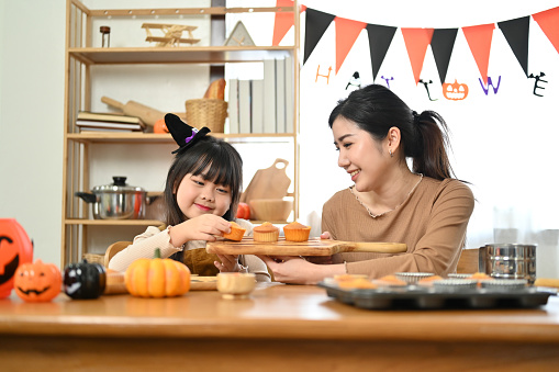 Pretty and joyful little Asian girl in fancy costume making Halloween cupcakes for the Halloween party with her mom.