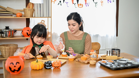 An adorable young Asian girl is concentrating on decorating her Halloween cupcake in the kitchen with her mom. Happy family concept