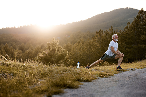 Happy mature athlete stretching while preparing for sports training in nature at sunset. Copy space.