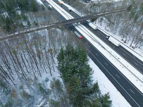 Highway through a snowy forest landscape seen from above with colorful cars and tracks driving on the black asphalt. The A50 highway through the Veluwe nature reserve is surrounded by snow covered pine trees.