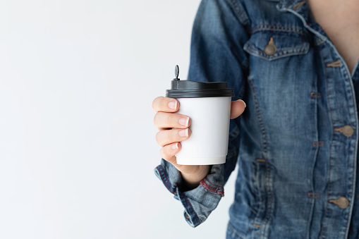 Unrecognizable caucasian female wearing a denim jacket top is showing a disposable cup in hand to camera.