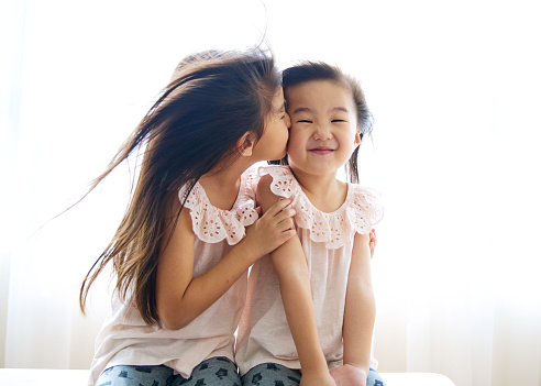 2 cute little lovely asian sisters sitting at home, while elder sister kiss on shy younger sister