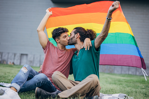 Male gay couple sitting on grass and holding gay flag