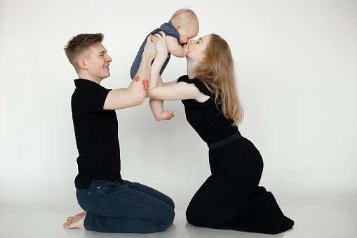 Portrait of young happy beautiful family in dark clothes kneeling with little plump grey-eyed baby infant on white background. Parents holding child high above heads, kissing. Family, having fun.