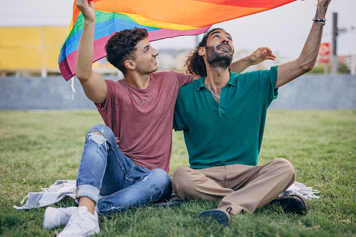 Male gay couple sitting on grass and holding gay flag