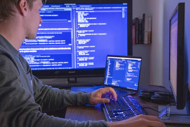 Young man uses multiple computers while coding, developing web site stock photo